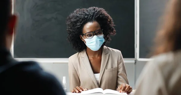 안경과 의료용 마스크를 착용 한 아프리카계 미국인젊은 여성 교사가 학교에서 테이블에 앉아 교과서를 읽고 가르치고 있습니다. 문학 수업이요. 학생들이나 학생들 앞에서의 여성 교육자. — 스톡 사진