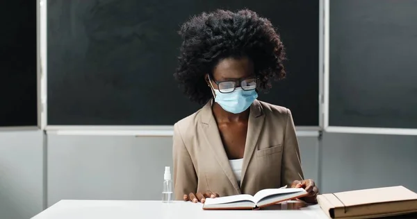Афроамериканська вчителька в окулярах і медичній масці сидить за столом у школі, читаючи підручник і викладаючи. Урок з літератури. Жінка-педагог ставить запитання студентам або учням. — стокове фото