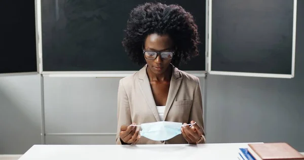 Retrato de una profesora afroamericana con gafas tomando máscara médica, sentada en la mesa en el aula de la escuela y sonriendo a la cámara. Profesora con protección. Concepto de Coronavirus. —  Fotos de Stock