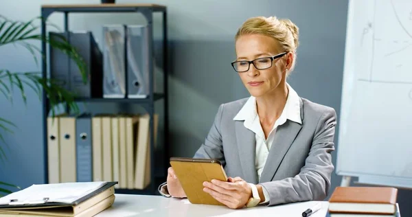Портрет кавказской предпринимательницы в очках, сидящей в офисе и использующей планшетное устройство, улыбающейся в камеру. Женщина в деловом стиле нажатия и прокрутки на гаджет-компьютер за столом в шкафу. — стоковое фото