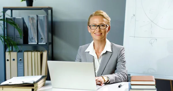 Портрет кавказской предпринимательницы в очках, сидящей в офисе и работающей на ноутбуке, улыбающейся в камеру. Женщина в деловом стиле печатает на клавиатуре компьютера за столом в шкафу. — стоковое фото