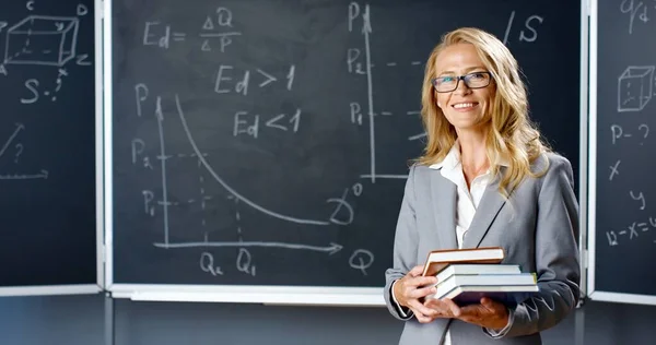 Porträt einer schönen kaukasischen Lehrerin, die im Klassenzimmer an Bord steht, in die Kamera lächelt und Schulbücher in der Hand hält. Dozentin mit Büchern an der Tafel mit mathematischen Formeln und Zeichnungen. — Stockfoto