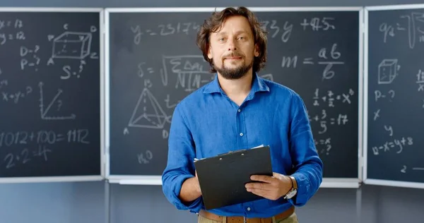 Retrato de un maestro caucásico de pie en el aula con una carpeta en las manos mirando a la cámara. Fórmulas matemáticas y leyes de fondo. Profesor matemático que trabaja en la escuela o la universidad. —  Fotos de Stock