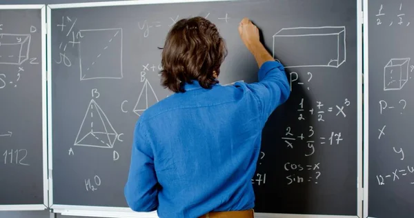 Back view Caucasian male lecturer writing math or physics formulas with chalks on blackboard. Man teacher working at school. Lesson of mathemathics. Mathematician. Geometry or algebra equation. Rear. Royalty Free Stock Images