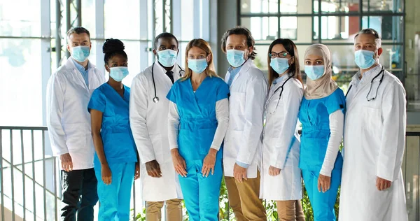 Mixed-races team of specialists, males and females doctors in hospital. International group of medics in medical masks. Protected workers. Multi ethnic physicians and nurses in uniforms in clinic. — Stock Photo, Image