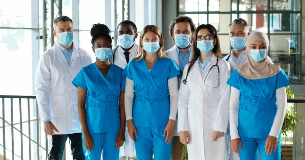 Mixed-races busy team of male and female doctors posing to camera and crossing hands in hospital. International group of medics in medical masks. Protected multi ethnic physicians and nurses in clinic — Stock Photo, Image