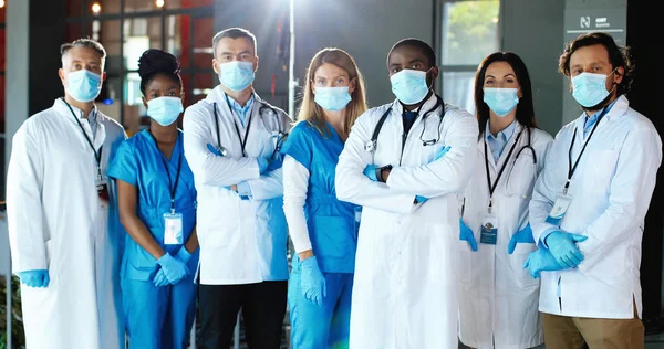 Multi ethnic women and men, doctors. International medics in medical masks. Protected healthcare workers. Mixed-races physicians and nurses looking at camera. Clinic team. Docs at work in hospital.