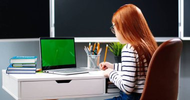 Uzun kızıl saçlı, tıbbi maskeli, ödev ödevlerini defter içinde yazan ve masa başında oturan hoş beyaz bir kız. Yeşil ekranlı dizüstü bilgisayar. Krom anahtar. Arka taraf. Okul hayatı.