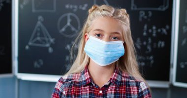 Sınıfta kameraya bakan, tıbbi maskeli, güzel, beyaz bir kızı yakından çek. Okulun kara tahtasında sarışın güzel bir kız. Coronavirus salgını araştırması.