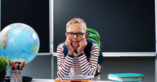 Blanke schattige tiener jongen met bril en met rugzak komt in de klas. Portret van een schooljongen die op een bureau leunt en glimlacht naar de camera. Het schoolleven. Het begrip "jeugd". — Stockfoto