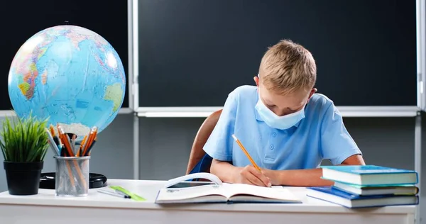 Pequeño chico adolescente caucásico en máscara médica sentado en el escritorio con el mundo en la escuela. Adolescente escolar haciendo tarea y ejercicio de escritura en copybook. Aprendizaje de los alumnos en el aula. Concepto de Coronavirus — Foto de Stock