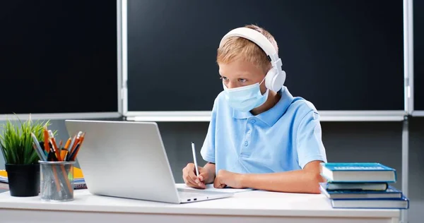 Pequeño chico adolescente caucásico con máscara médica y auriculares sentados en el escritorio de la escuela. Adolescente escolar haciendo tarea y ejercicio de escritura en copybook delante de la computadora portátil. Concepto Covid-19. — Foto de Stock