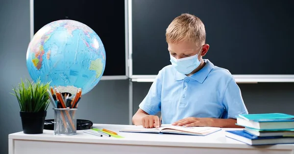 의료용 마스크를 쓰고 책상에 앉아 있는 작은 백인 십 대 소년이 학교에서 지구와 함께 앉아 있습니다. 교과서를 읽고 손가락으로 따라가는 10 대 남학생. 교실에서 배울 수있습니다. 코로나 바이러스의 개념. — 스톡 사진