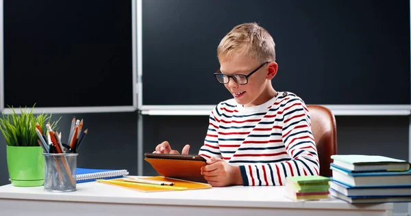 Biały mały nastolatek w masce medycznej siedzi przy biurku w szkole i bawi się na tablecie, stukając i przewijając na ekranie. Mały uczeń korzystający z komputera z gadżetami i smsów. Koncepcja pandemii. — Zdjęcie stockowe
