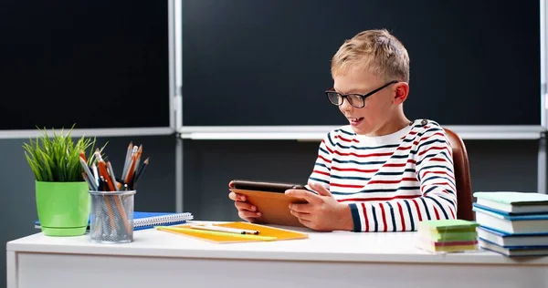 Petit adolescent caucasien en masque médical assis au bureau à l'école et jouant sur un appareil tablette, tapotant et défilant à l'écran. Petit écolier utilisant un ordinateur gadget et textos. Concept pandémique. — Photo