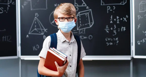 Porträtt av söt liten vit pojke i glasögon, medicinsk mask och med ryggsäck stående i klassrummet, tittar på kameran. En liten skolpojke i glasögon i skolan. Begreppet pandemi med coronavirus. — Stockfoto