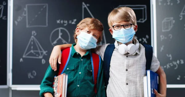 Porträtt av söta små kaukasiska pojkar bästa vänner i medicinska masker, med ryggsäckar och läroböcker står i klassrummet vid svarta tavlan. Små skolpojkar kramas i skolan. Coronaviruspandemi. — Stockfoto