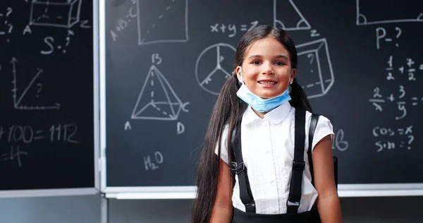 Retrato de linda niña caucásica feliz pequeña con mochila de pie en el aula, mirando a la cámara y quitándose la máscara médica. Una colegiala hispana sonriendo en la escuela. Coronavirus. —  Fotos de Stock