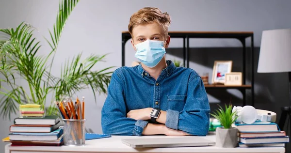 Retrato de adolescente caucásico con máscara médica sentado en el escritorio con libros y libros de texto y mirando a la cámara. Escolar adolescente. Concepto de tarea. Vida escolar. Concepto pandémico del Coronavirus. —  Fotos de Stock