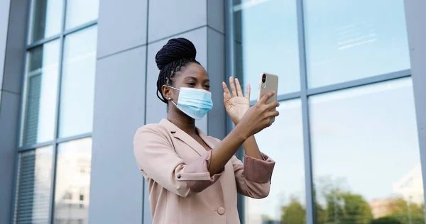Lykkelig afroamerikansk ung kvinne med medisinsk maske, med videokapsel på smarttelefon utendørs i forretningsbygg. Munter vakker kvinne som snakker og videokhatter via nettkamera på mobil. – stockfoto