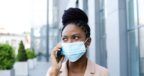 Afro-americano jovem bela mulher alegre em máscara médica falando no telefone celular e rindo do lado de fora no centro de negócios. Empresária feliz na proteção respiratória falando no celular. — Fotografia de Stock