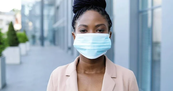 Retrato de una joven afroamericana hermosa mujer en chaqueta rosa quitándose la máscara médica y sonriendo a la cámara al aire libre. Atractiva mujer de negocios alegre en la calle cerca del centro de negocios en cuarentena. —  Fotos de Stock