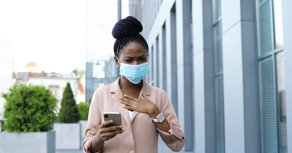 Jovem mulher de negócios afro-americana doente em máscara médica tossir e usar smartphone ao ar livre. Mulher doente com sintoma de coronavírus na rua perto do centro de negócios. Mulher não saudável com telefone celular. — Fotografia de Stock