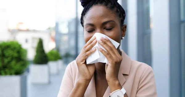 Ung, syk, afroamerikansk forretningskvinne som hoster og nyser utendørs. Syk kvinne med coronavirus symptom på gaten nær forretningssenteret. Usunne kvinner nyser og hoster. Kokt begrep. – stockfoto