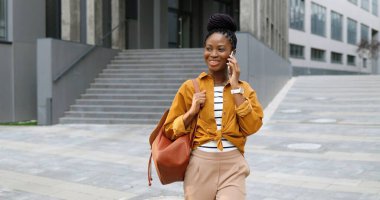 Afro-Amerikan güzel, mutlu, gülümseyen, cep telefonuyla konuşan ve dışarıda yürüyen bir kadın. Neşeli bayan cep telefonuyla konuşuyor ve geziniyor. Telefon görüşmesi.