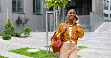 Afro-Amerikan güzel, mutlu, gülümseyen, cep telefonuyla konuşan ve dışarıda yürüyen bir kadın. Neşeli bayan cep telefonuyla konuşuyor ve geziniyor. Telefon görüşmesi.
