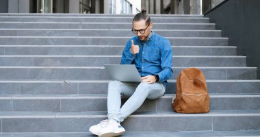 Beyaz tenli gözlüklü bir genç dışarıda oturuyor ve web kamerasıyla dizüstü bilgisayarında konuşuyor. Erkek öğrenci ya da serbest çalışan biri sokakta görüntülü sohbet ediyor ve düşünüyor. Görüntülü arama. Başparmağımı bırakıyorum..