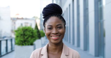 Pembe ceketli güzel Afro-Amerikan bir kadının portresi. Kameraya bakıyor ve dışarıda neşeyle gülümsüyor. Çekici, mutlu, gülümseyen iş kadını iş merkezinin yanındaki sokakta..