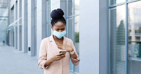 Ung sjuk afroamerikansk affärskvinna i medicinsk mask hosta, gå och använda smartphone utomhus. Sjuk kvinna med coronavirus symptom på gatan. Ohälsosam kvinna med mobiltelefon kliva. — Stockfoto