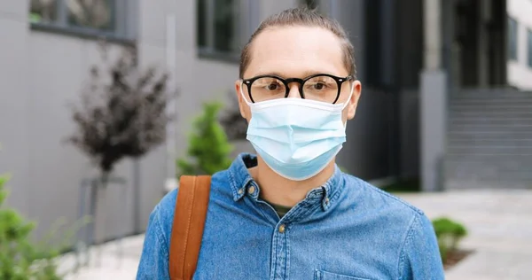 Retrato de un joven caucásico guapo con gafas quitándose la máscara médica y sonriendo a la cámara al aire libre. Hombre alegre en la calle en cuarentena. Acercándonos. Tipo con anteojos y protección contra virus. —  Fotos de Stock