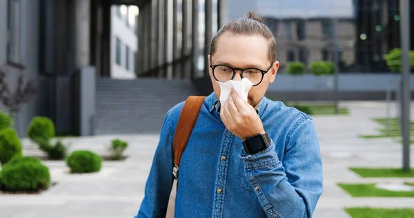 Young ill Caucasian man coughing and sneezing in napkin outdoor. Sick male with coronavirus symptom at street near business center. Unhealthy guy sneeze and cough. Covid pandemic concept.