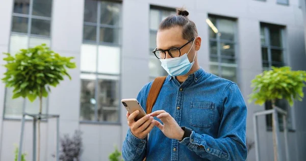 안경을 쓰고 의료 마스크를 쓴 백인 청년이 시내 거리에서 핸드폰으로 도청을 하고 있습니다. 야외에 있는 스마트폰으로 문자 메시지를 보내는 모습입니다. 개념을 사용하는 가젯. — 스톡 사진