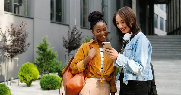 Ženy ze smíšených ras si povídají a dívají se na něco na mobilu na ulici ve městě. Krásné multietnické mladé ženy chatují a používají smartphone. Veselí přátelé pomlouvají. Gossips. — Stock fotografie