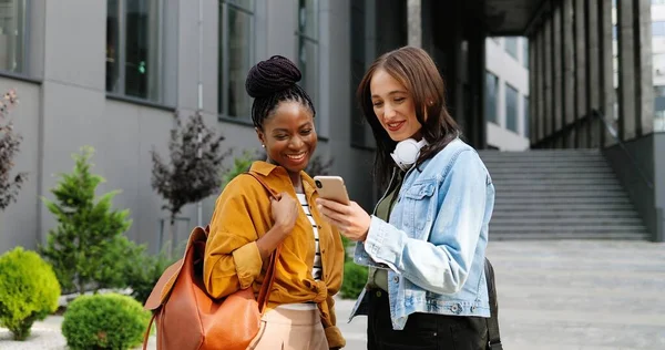 Blandade raser kvinnor pratar och titta på något på mobiltelefon på gatan i staden. Vackra multietniska unga kvinnor chattar och använder smartphone. Glada vänner som skvallrar. Skvaller. — Stockfoto