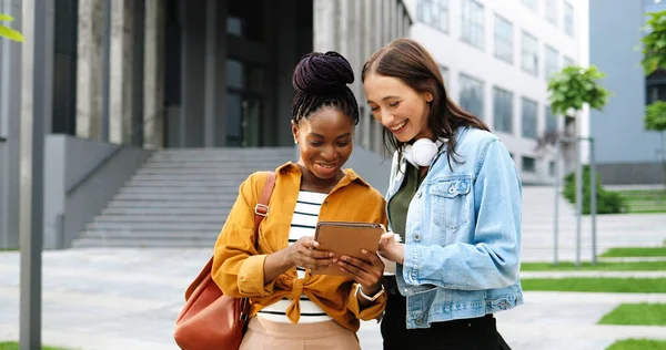 Mestiços fêmeas falando e assistindo algo no dispositivo tablet na rua na cidade. Mulheres jovens multi étnicas bonitas conversando e usando o computador gadget. Amigos alegres a coscuvilhar. Fofos. — Fotografia de Stock