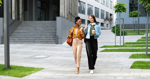 Mixed-races young pretty females, best friends talking cheerfully and walking with cups of coffee to-go and at city street. Multi ethnic beautiful happy women students strolling outside with drinks.