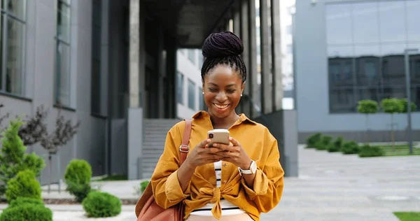 아프리카계 미국인을 기쁘게 하는 젊은 여성이 스마트폰으로 두드리거나 스크롤을 하며 시가지에 서 있습니다. 아름다운 여성이 핸드폰으로 문자 메시지를 보내고 웃고 있습니다. 밖. — 스톡 사진