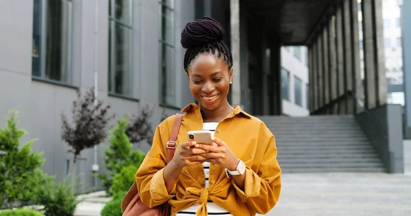 Afro-americano alegre jovem mulher elegante tocando ou rolando no smartphone e de pé na rua da cidade. Linda mensagem de texto feminino feliz no telefone celular e sorrindo. Fora. — Fotografia de Stock