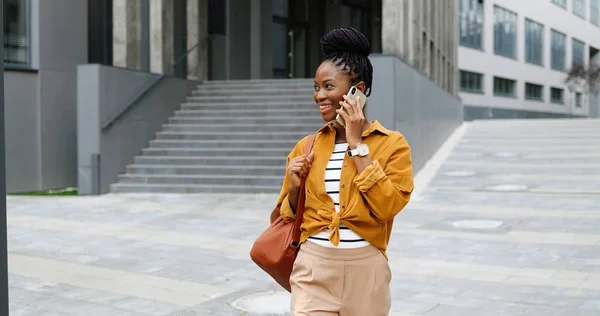 Afrikansk amerikan vacker glad leende kvinna talar på mobiltelefon och går utanför. Glada kvinna talar på mobiltelefon och promenader. Telefonsamtal. — Stockfoto