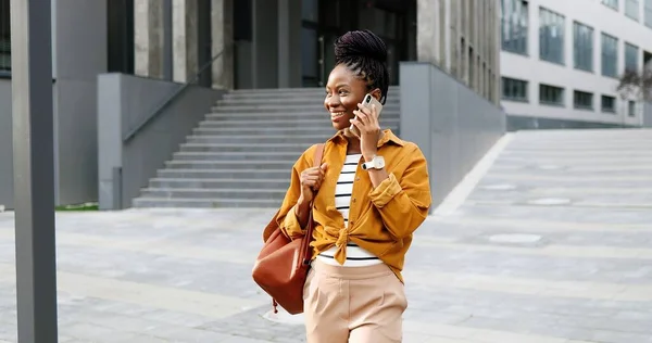 Afro-americano bela feliz sorriu mulher falando no telefone celular e andando fora. Mulher alegre falando no celular e passeando. Conversa telefónica. — Fotografia de Stock