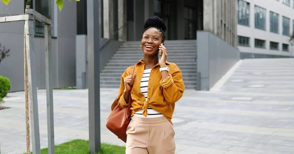 Afro-americano bela feliz sorriu mulher falando no telefone celular e andando fora. Mulher alegre falando no celular e passeando. Conversa telefónica. — Fotografia de Stock