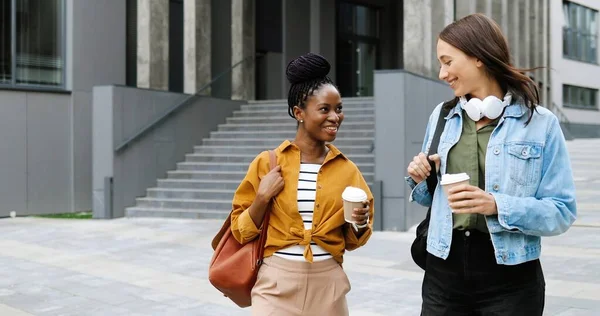 Mixed-races young pretty females best friends talking cheerfully and walking with cups of coffee to-go and at city street. Multi ethnic stylish happy women students strolling outside with hot drinks