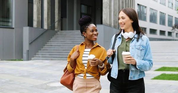 Mixed-races young pretty females best friends talking cheerfully and walking with cups of coffee to-go and at city street. Multi ethnic stylish happy women students strolling outside with hot drinks