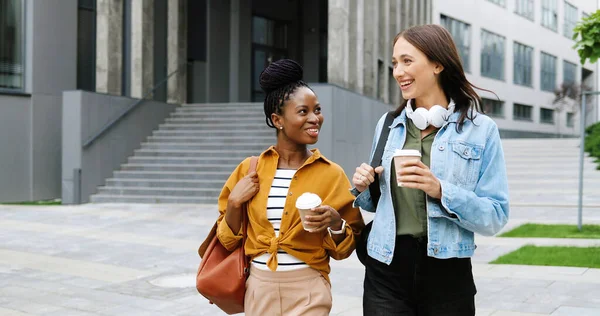 Blandade raser unga vackra kvinnor bästa vänner talar glatt och promenader med koppar kaffe att gå och på stadens gata. Multi etniska snygga glada kvinnor studenter promenader utanför med varma drycker — Stockfoto