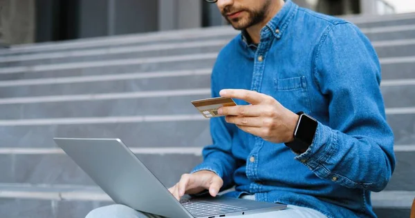 Caucasian young man in glasses and blue jeans shirt using credit card and shopping online on laptop. Good-looking male in eyeglasses typing on computer and buying in internet. Buyer at street. — Stock Photo, Image