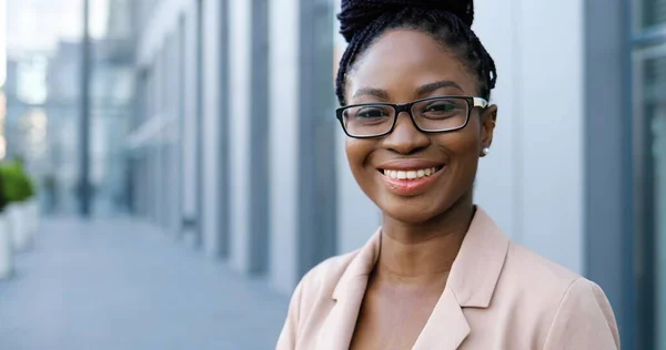 Πορτρέτο της νεαρής Αφροαμερικανής όμορφη γυναίκα σε ροζ σακάκι και γυαλιά που αναζητούν την κάμερα και χαμογελώντας χαρούμενα σε εξωτερικούς χώρους. Ελκυστική σοβαρή επιχειρηματίας στο δρόμο κοντά στο επιχειρηματικό κέντρο. — Φωτογραφία Αρχείου